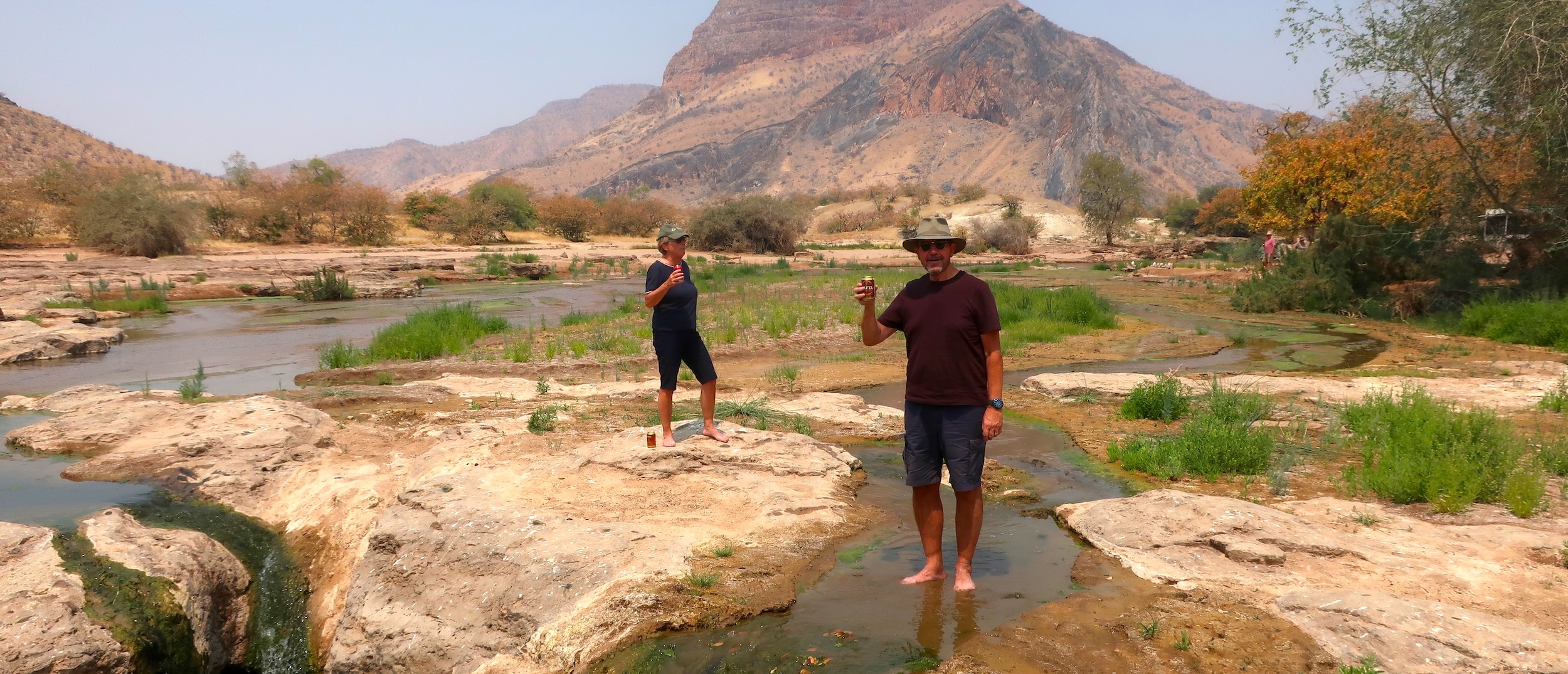 Two tourists pose with drinks while standing in a shallow river.