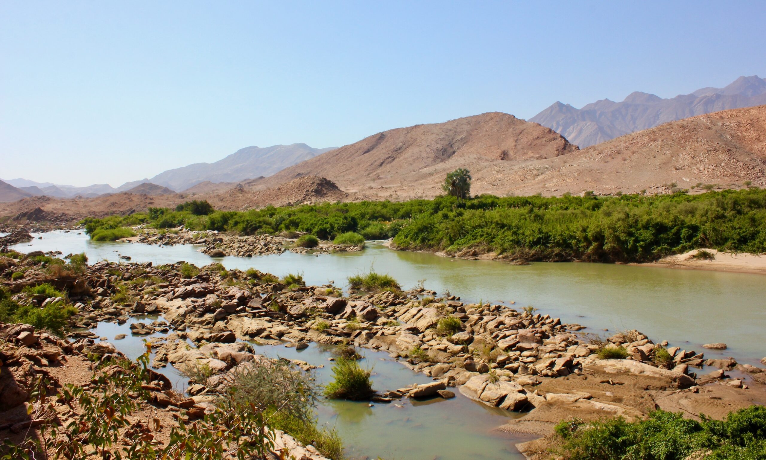 Rocky shoreline beside a river with red hills in the background.